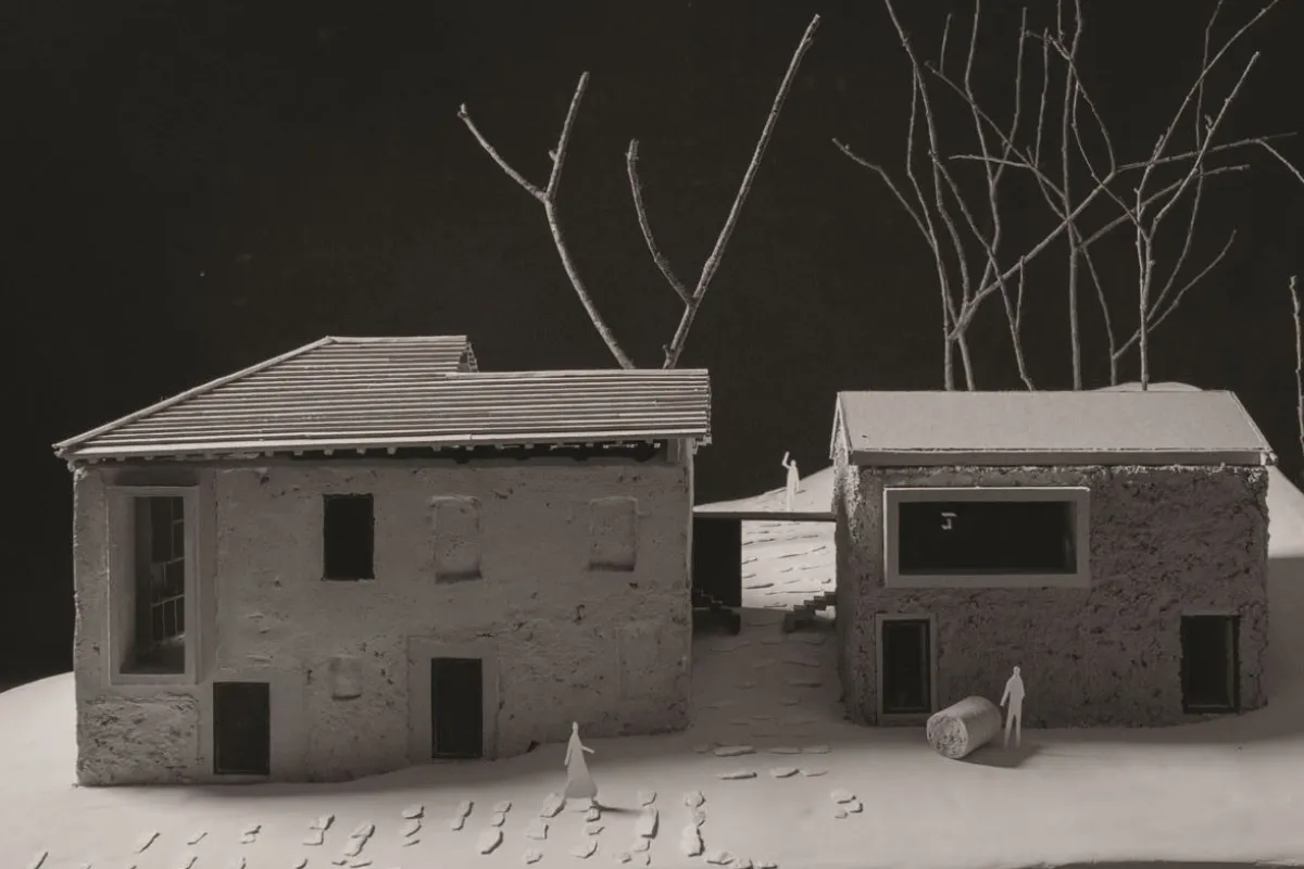 Modell eines architektonischen Projekts mit zwei Gebäuden, umgeben von kahlen Bäumen. Das linke Gebäude hat ein geneigtes Dach, während das rechte ein Flachdach aufweist. Kleine Figuren und Fußspuren sind auf dem Boden sichtbar, was eine menschliche Präsenz andeutet.