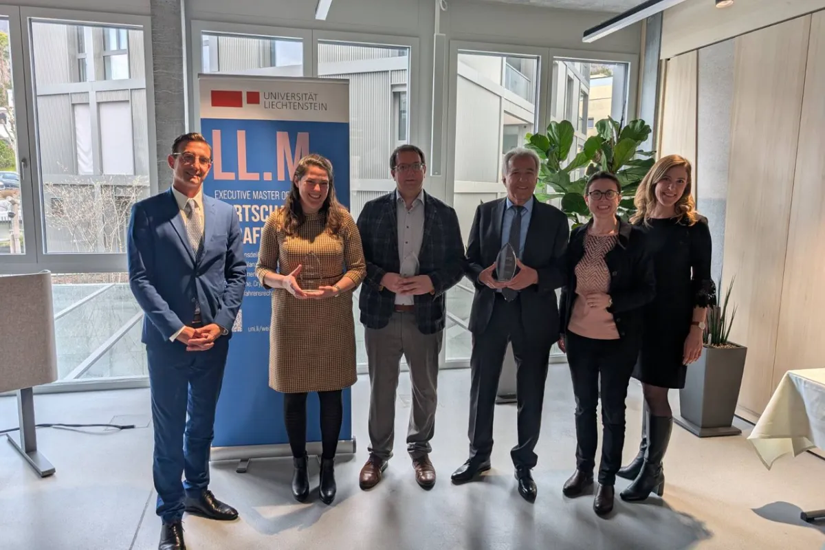 Sechs Personen stehen in einem modernen Raum vor einem Banner der Universität Liechtenstein. Zwei Personen halten Glastrophäen. Die Gruppe lächelt in die Kamera. Im Hintergrund sind große Fenster und Pflanzen zu sehen.