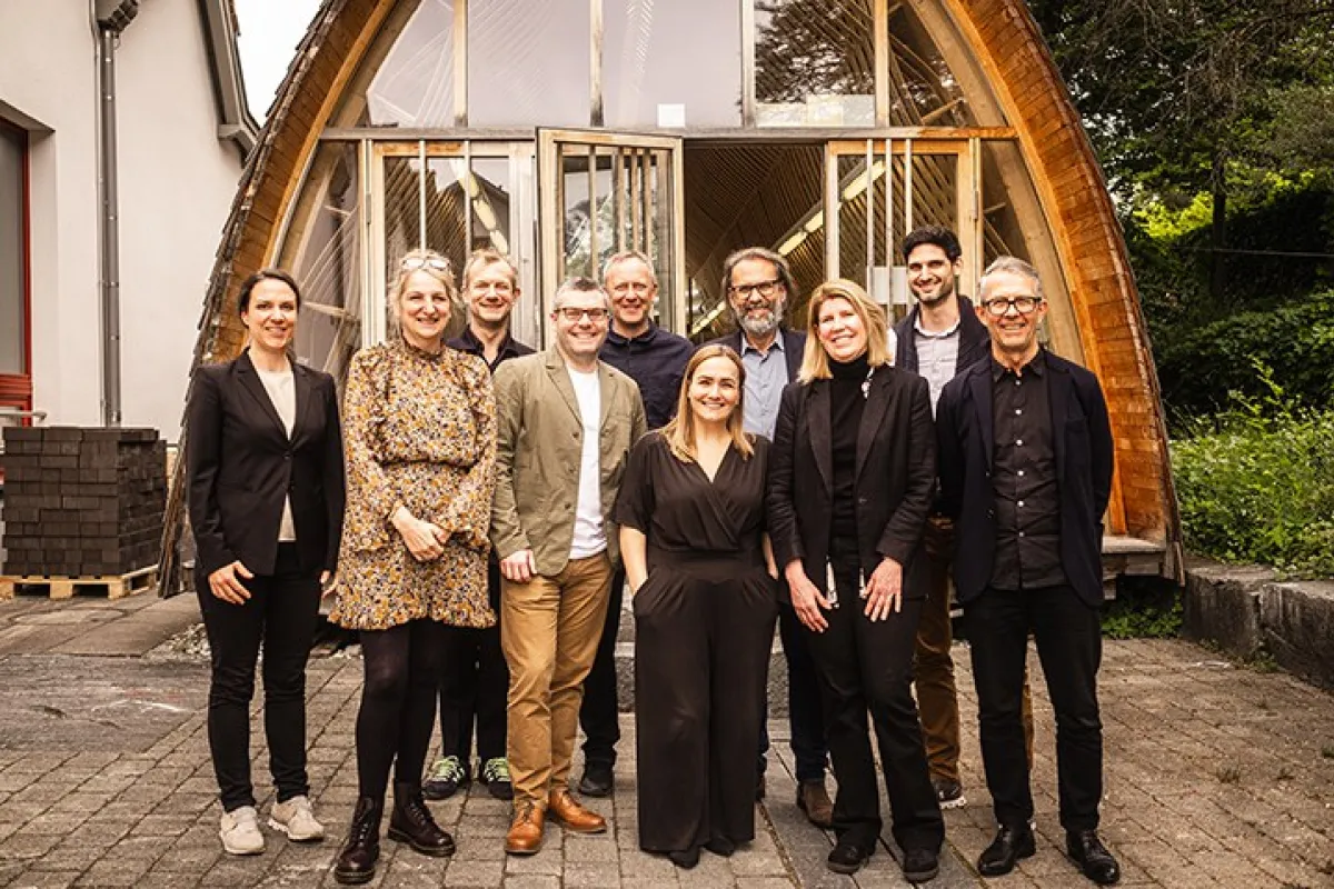 Eine Gruppe von zehn Personen steht lächelnd vor einem modernen Holzgebäude mit großen Fenstern. Die Szene vermittelt eine freundliche und professionelle Atmosphäre.