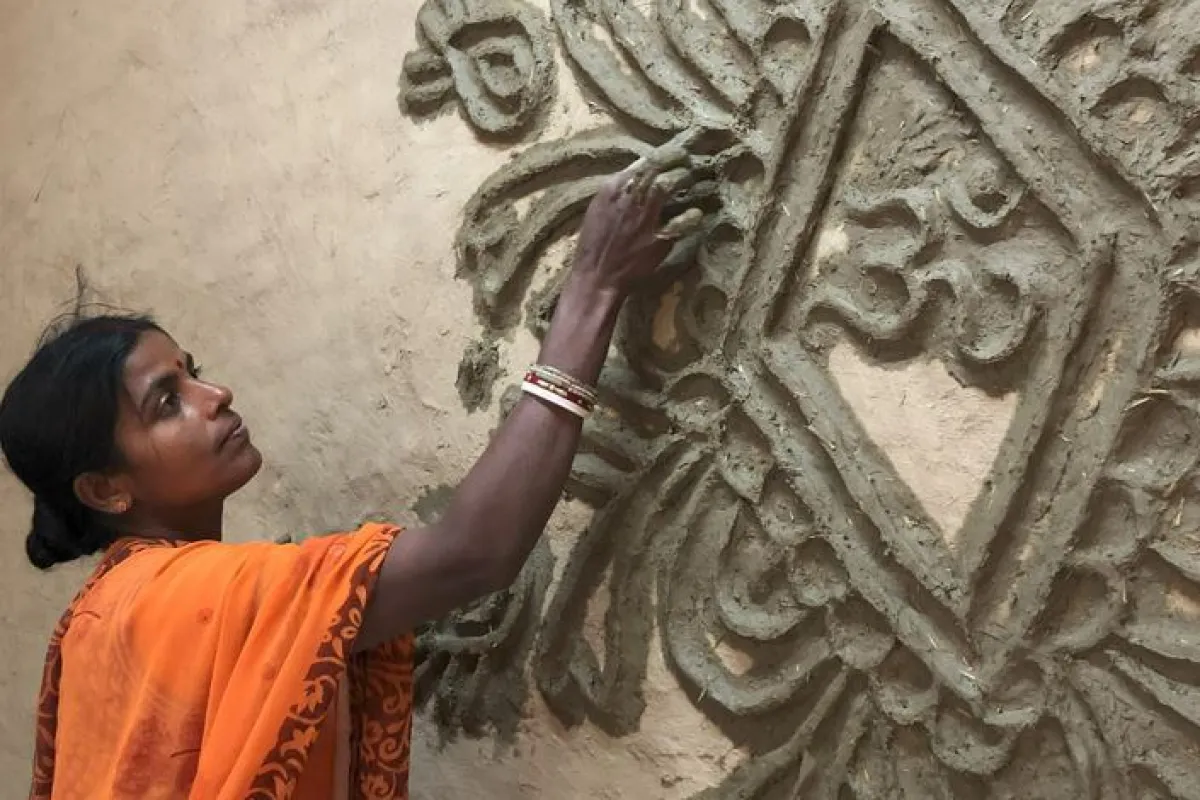 Eine Frau in einem orangefarbenen Sari verziert eine Wand mit traditionellen Tonmustern. Sie arbeitet konzentriert an einem kunstvollen Relief.