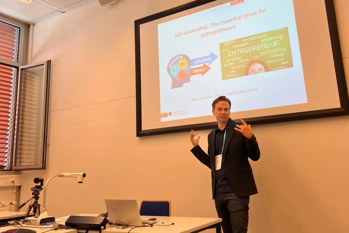 Ein Vortragender präsentiert in einem Seminarraum ein Paper über Selbstführung für Unternehmer. Im Hintergrund ist eine Leinwand mit einer Präsentationsfolie zu sehen. Tische mit Laptops und Kamera sind im Vordergrund.