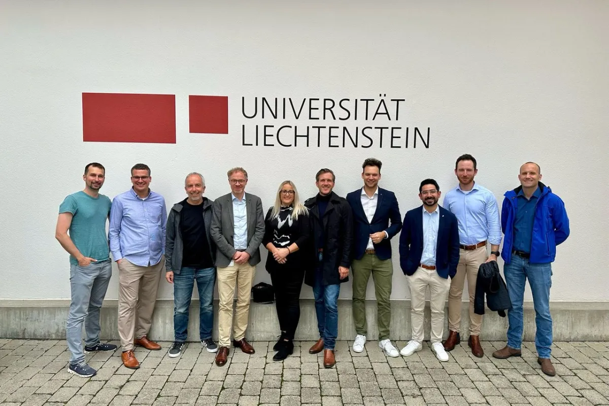 Gruppe von zehn Personen steht lächelnd vor einer Wand mit dem Logo der Universität Liechtenstein. Sie tragen unterschiedliche Freizeit- und Businesskleidung.