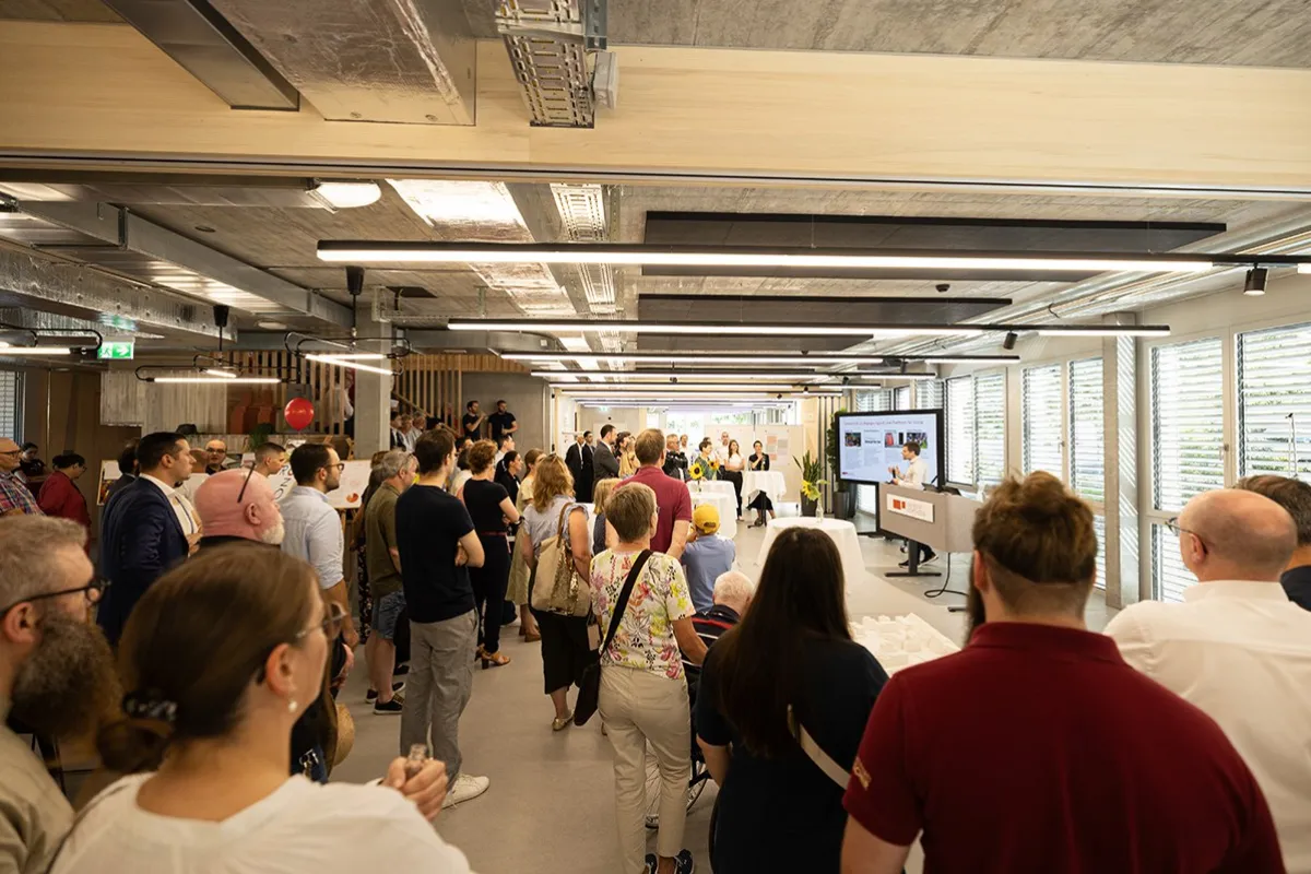 Besuchergruppe beim Tag der offenen Tür an der Universität Liechtenstein, neuer Standort Ebaholz. Menschen stehen in einem modernen Raum und hören einer Präsentation zu. Im Hintergrund sind große Fenster und eine Leinwand zu sehen.