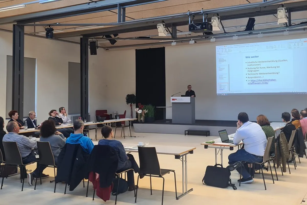 Eine Gruppe von etwa 20 Personen sitzt in einem Auditorium der Universität Liechtenstein, während ein Vortragender an einem Rednerpult spricht. Auf einer großen Leinwand ist eine Präsentation mit dem Titel "Wie weiter" zu sehen.