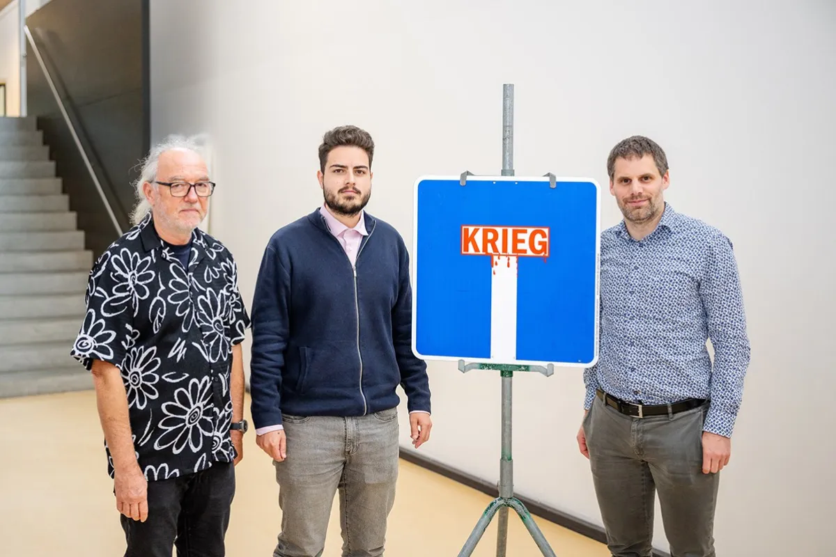 Drei Männer stehen neben einem blauen Verkehrsschild mit der Aufschrift "Krieg", das symbolisch eine Sackgasse darstellt. Das Bild wurde bei der Veranstaltung "Verkehrsschilder der Gerechtigkeit" an der Universität Liechtenstein aufgenommen.
