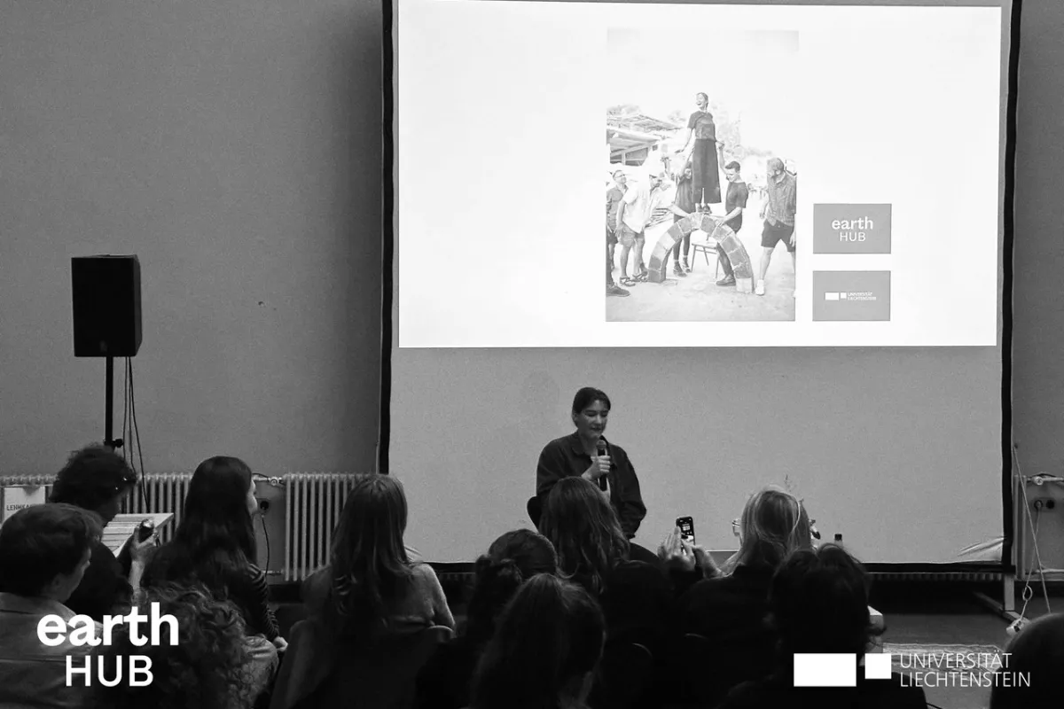 Eine Vortragende steht vor einem Publikum und präsentiert. Im Hintergrund ist eine Leinwand zu sehen. Das Bild ist in schwarz-weiss.