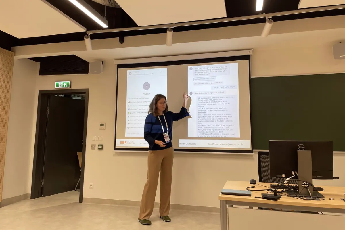 Eine Person präsentiert in einem modernen Seminarraum vor einer Leinwand mit zwei projizierten Dokumenten. Im Vordergrund steht ein Tisch mit einem Computerbildschirm.