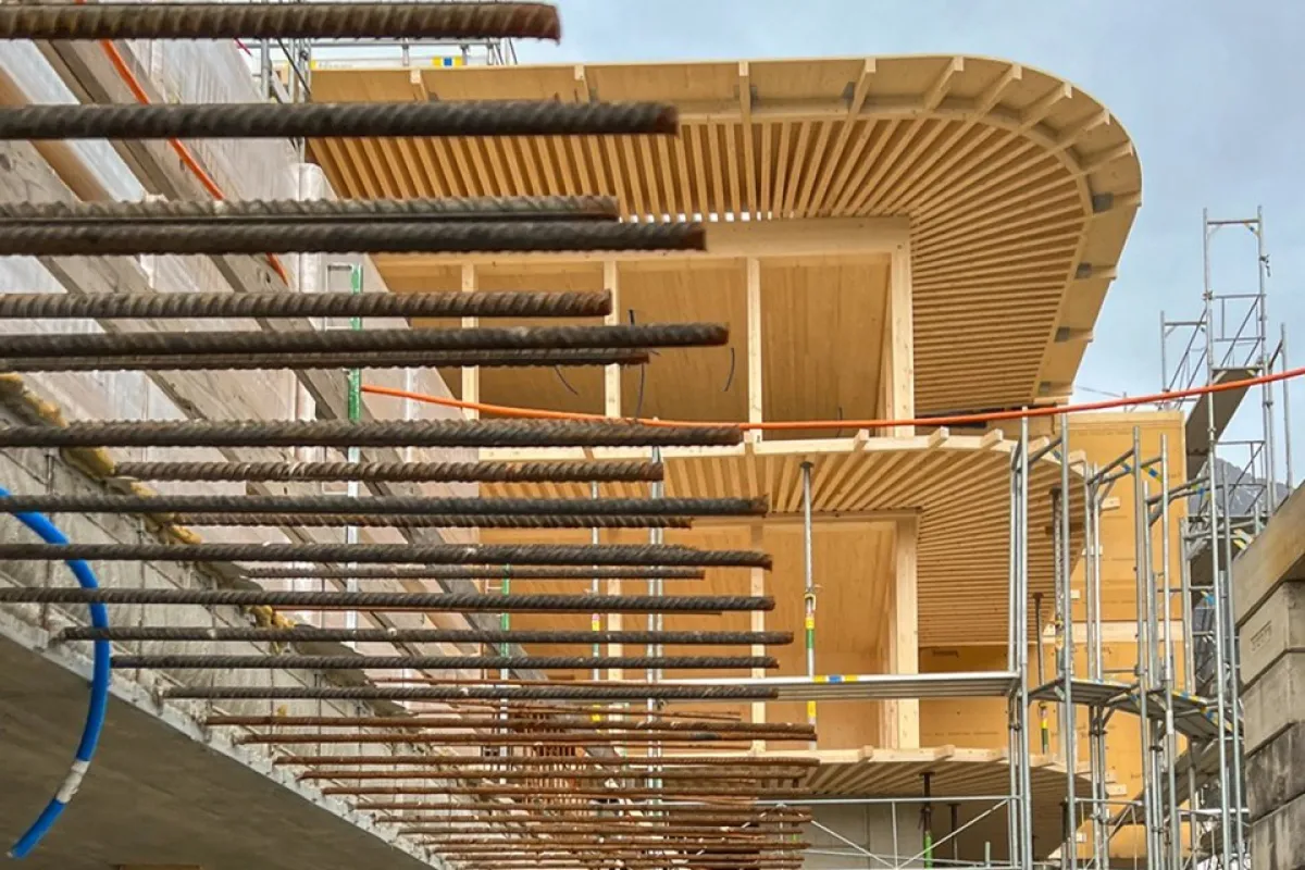 Baustelle eines modernen Gebäudes mit sichtbaren Stahlträgern und Holzverkleidung, umgeben von Gerüsten, unter bewölktem Himmel.