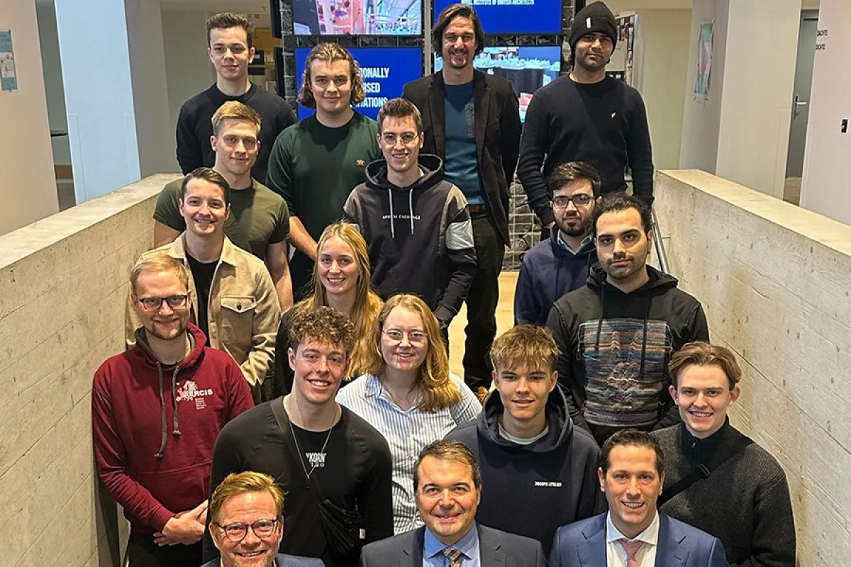 Gruppe von Studierenden und Lehrenden steht auf einer Treppe in einem modernen Gebäude der Universität Liechtenstein, lächelnd in die Kamera. Im Hintergrund sind Bildschirme und eine Steinwand zu sehen.