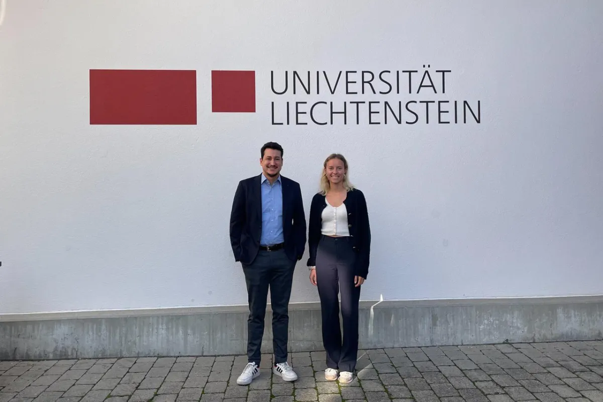 Zwei Personen stehen vor einer Wand mit dem Logo der Universität Liechtenstein. Sie lächeln in die Kamera.