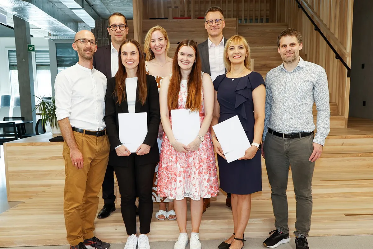 Eine Gruppe von acht Personen steht lächelnd in einem modernen Innenraum. Drei Personen in der vorderen Reihe halten Zertifikate in den Händen. Die Umgebung wirkt hell und einladend mit Holzelementen und Pflanzen.