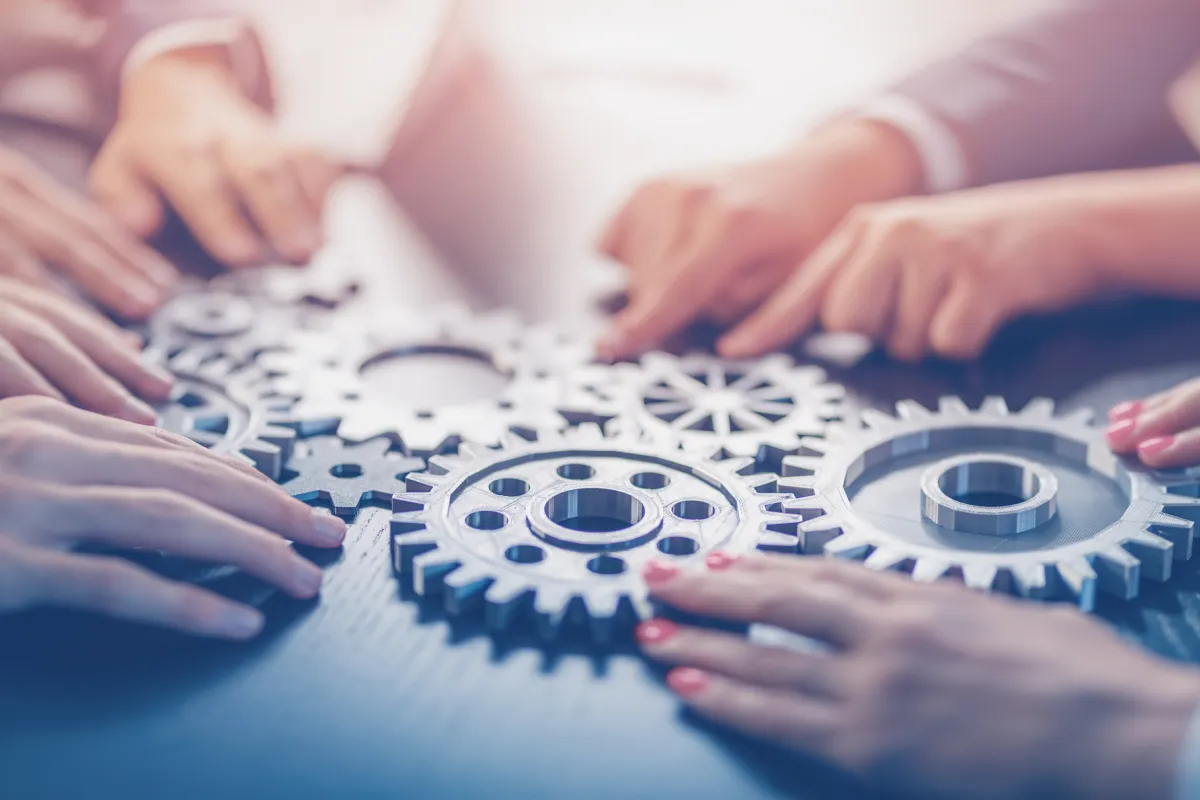 Mehrere Hände halten verzahnte Zahnräder als Symbol für Teamarbeit und Kooperation.