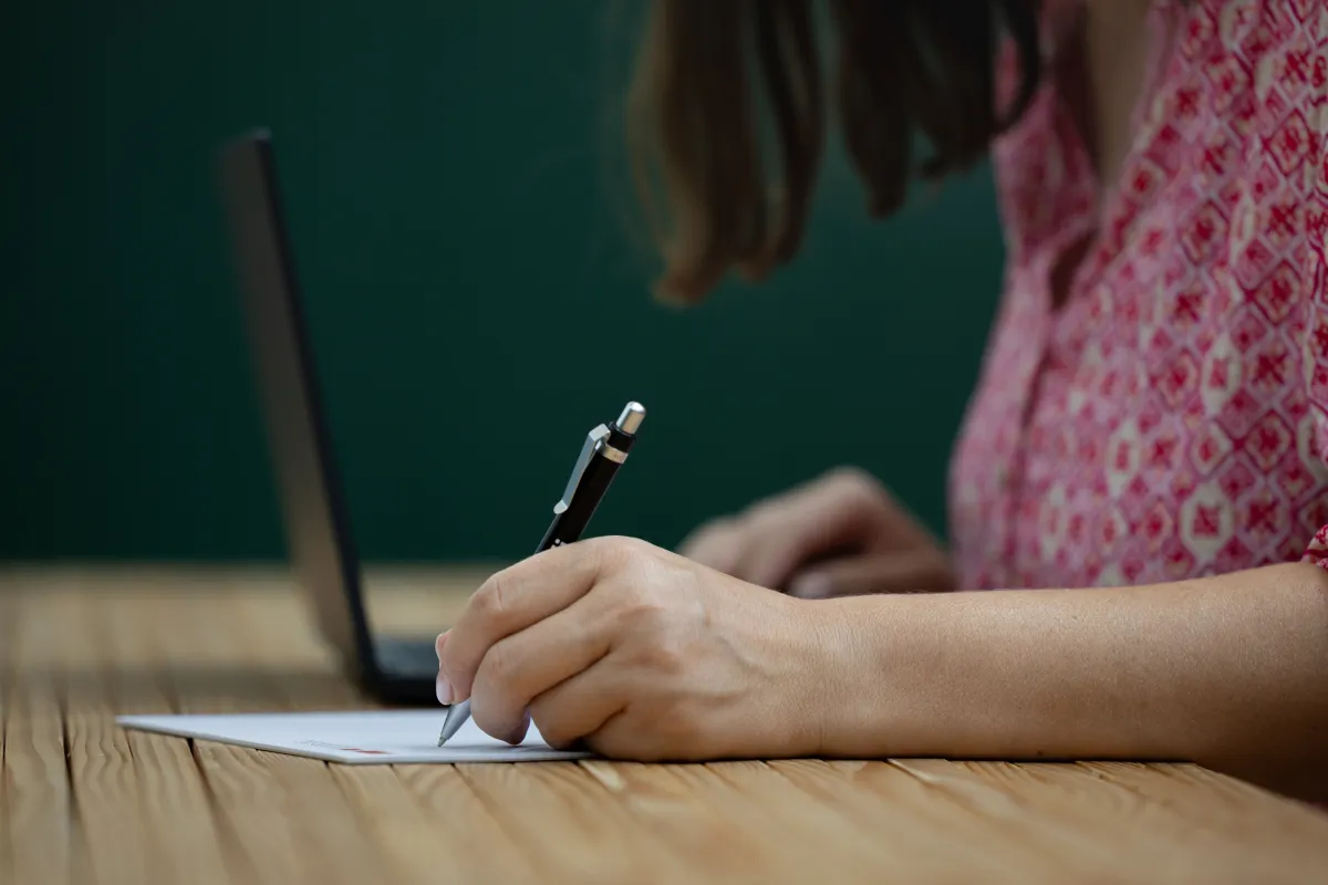 Person schreibt mit Stift auf Papier vor Laptop im Arbeitsraum der Universität Liechtenstein.