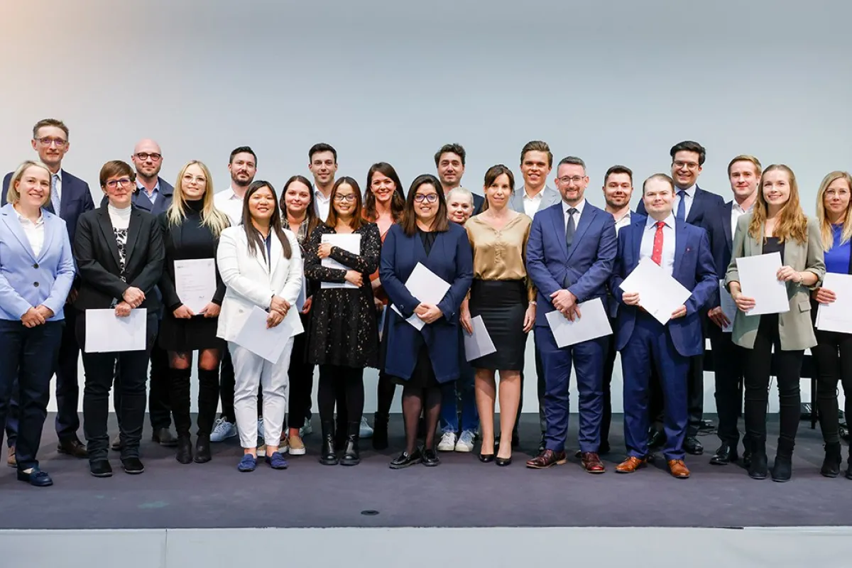Eine Gruppe von Absolventinnen und Absolventen steht auf einer Bühne und hält Zertifikate in den Händen. Sie lächeln in die Kamera. Die Personen sind formell gekleidet, und im Hintergrund ist eine neutrale Wand zu sehen.