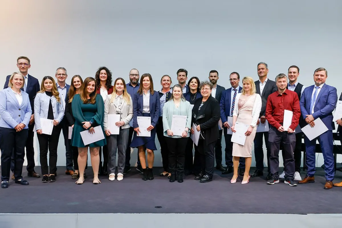 Eine Gruppe von Absolventinnen und Absolventen steht auf einer Bühne und hält Zertifikate in den Händen. Sie lächeln in die Kamera. Die Personen sind formell gekleidet, und im Hintergrund ist eine neutrale Wand zu sehen.