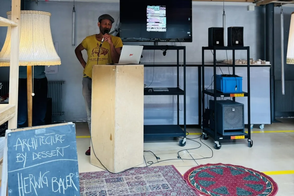 Ein Mann steht an einem Rednerpult in einem Raum mit Holzdach. Neben ihm ist ein Bildschirm mit einer Präsentation und ein Schild mit der Aufschrift „Architecture by Dessert, Herwig Bauer, Poolbarfestival“. Der Raum ist mit Teppichen und Stehlampen dekoriert.