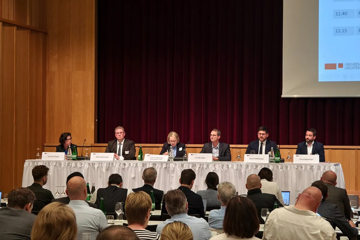 Ein Podium mit sechs Personen sitzt vor einem Publikum in einem Konferenzsaal. Auf dem Tisch stehen Namensschilder und Wasserflaschen. Im Hintergrund ist ein roter Vorhang und ein Teil einer Präsentationsleinwand zu sehen.