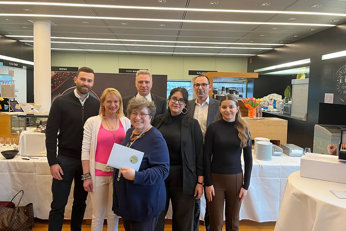 Eine Gruppe von sieben Personen steht lächelnd in einem modernen Raum. Eine Person hält ein Zertifikat in den Händen. Im Hintergrund sind Tische mit Geschirr und Dekorationen zu sehen.