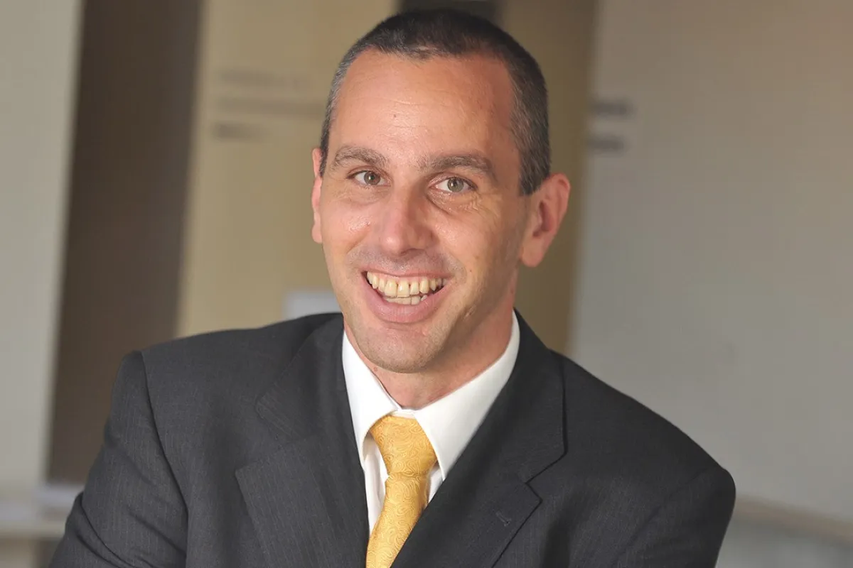 Porträt eines lächelnden Mannes in einem dunklen Anzug mit gelber Krawatte, vor einem neutralen Hintergrund.