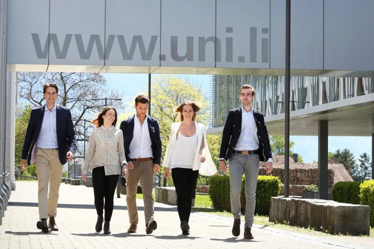 Eine Gruppe von fünf Personen geht lächelnd auf einem sonnigen Campusweg der Universität Liechtenstein entlang, mit der URL "www.uni.li" im Hintergrund.