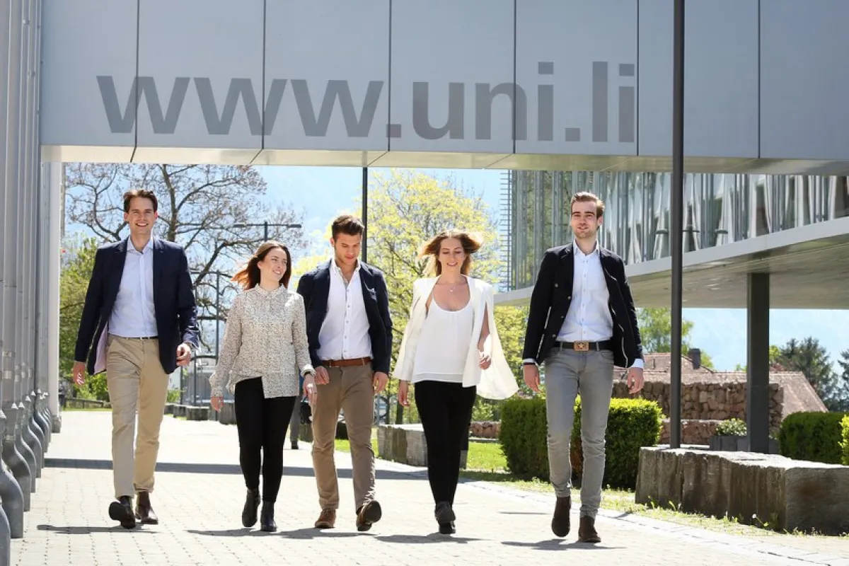Eine Gruppe von fünf Personen geht lächelnd auf einem sonnigen Campusweg der Universität Liechtenstein entlang, im Hintergrund moderne Architektur und Berge.