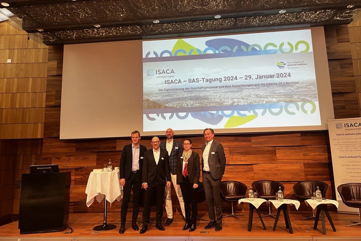 Gruppe von fünf Personen steht auf einer Bühne vor einer Leinwand mit dem Titel der ISACA-IIAS-Tagung 2024 in Zürich. Im Hintergrund sind Stühle und ein Tisch mit Wasserflaschen zu sehen.