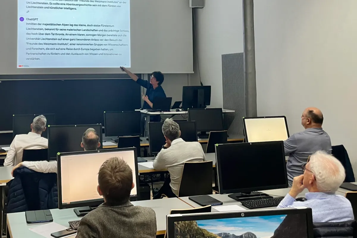 Ein Dozent hält einen interaktiven Workshop an der Universität Liechtenstein. Er zeigt auf eine Präsentation über künstliche Intelligenz, während die Teilnehmenden an Computern sitzen und aufmerksam zuhören.