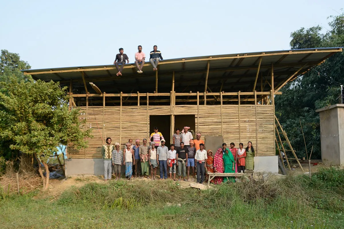 Eine Gruppe von Menschen steht vor einem im Bau befindlichen Gebäude aus Bambus in Bahuarwa, Indien. Drei Personen sitzen auf dem Dach. Das Bild zeigt den Fortschritt eines Schulbauprojekts, das von der Universität Liechtenstein unterstützt wird.