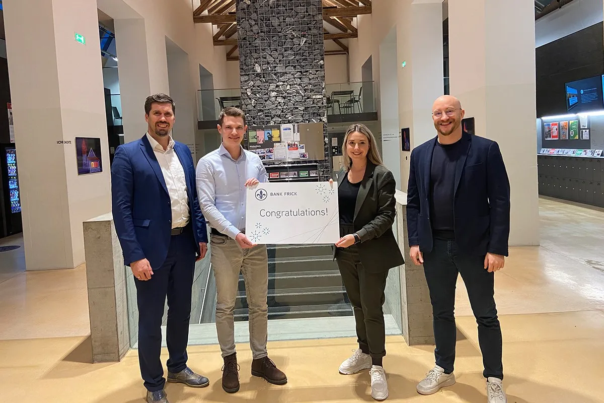 Vier Personen stehen in einem modernen Gebäude und halten ein Schild mit der Aufschrift "Congratulations!" von Bank Frick. Sie feiern die Vergabe eines Stipendiums für den Zertifikatsstudiengang Blockchain und FinTech an der Universität Liechtenstein.