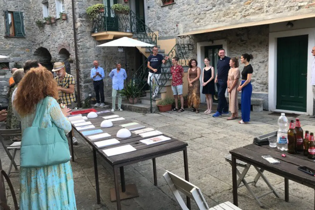 Eine Gruppe von Menschen versammelt sich in einem historischen Innenhof eines Dorfes. Auf Tischen liegen Dokumente und Objekte aus. Die Szene zeigt eine Präsentation oder Diskussion im Freien, umgeben von alten Steinmauern und Pflanzen.