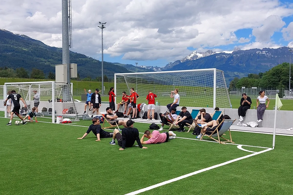 Teilnehmende des Völkerballturniers entspannen sich auf einem Sportplatz vor einer beeindruckenden Bergkulisse. Einige sitzen auf Liegestühlen, während andere am Spielfeldrand stehen. Im Hintergrund sind Fußballtore und Spieler in Sportkleidung zu sehen.