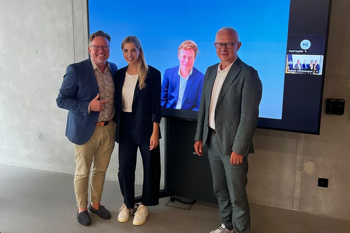 Drei Personen stehen vor einem Bildschirm, der zwei weitere Personen in einem virtuellen Meeting zeigt. Die Szene befindet sich in einem modernen Raum mit Betonwänden.