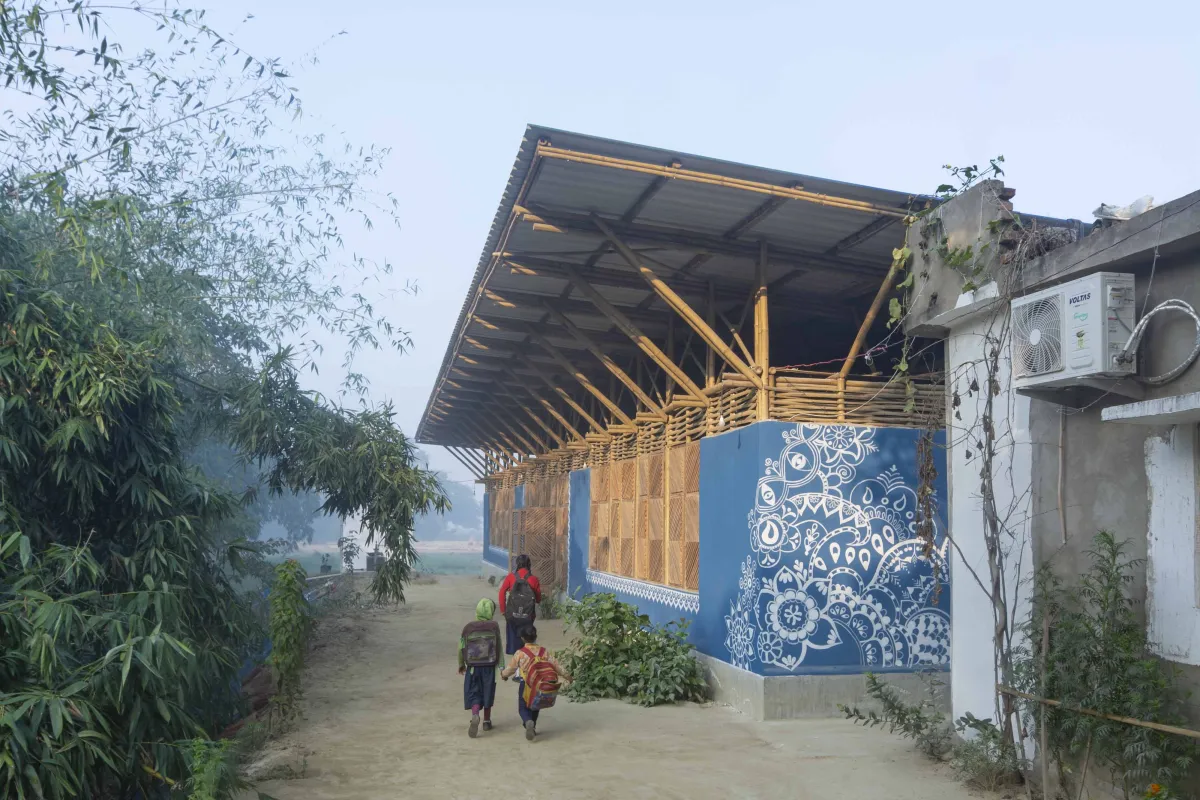 Kinder gehen auf einem sandigen Weg an einem blau bemalten Bambusgebäude mit weißem Ornamentmuster vorbei.