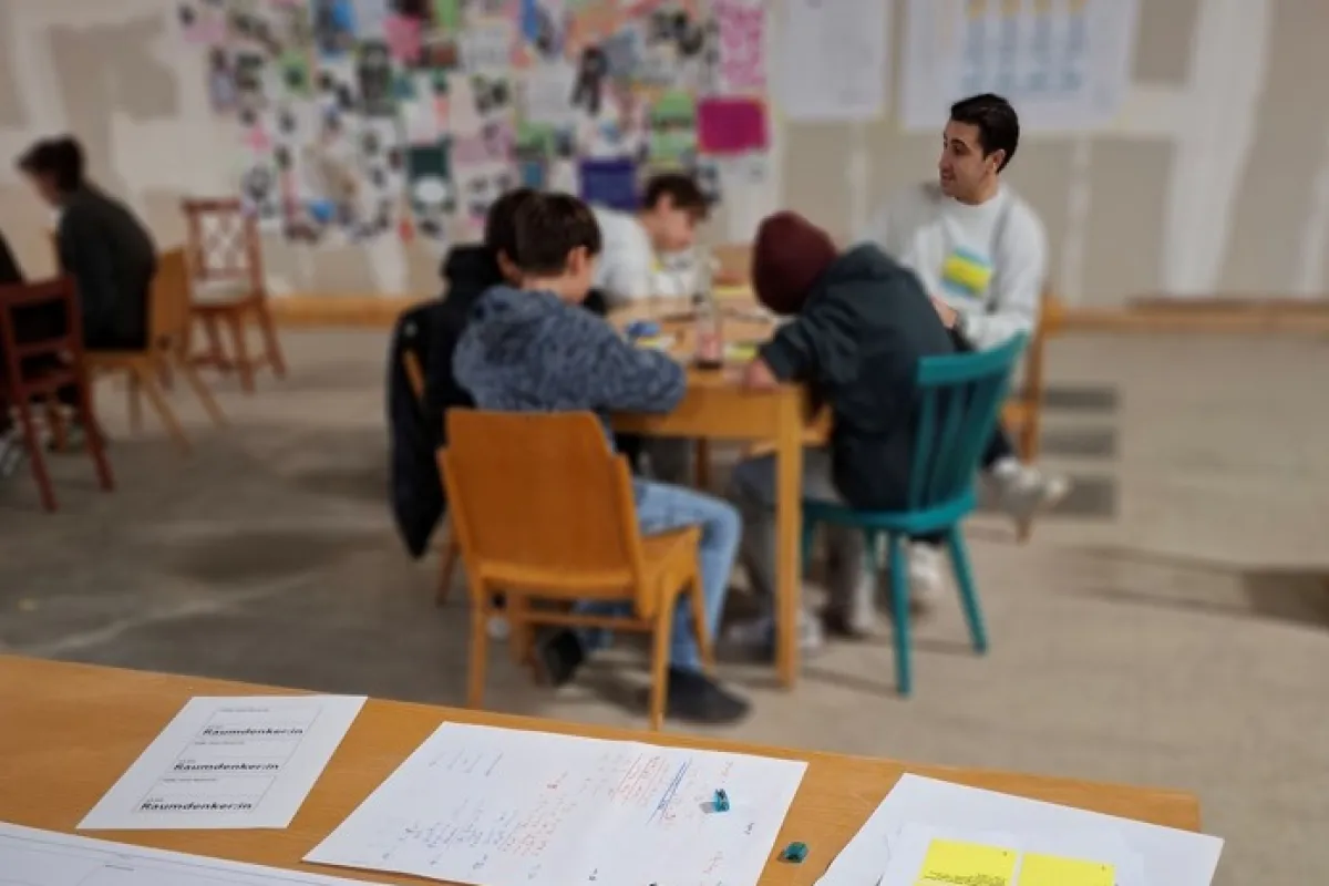 Blick auf einen Schreibtisch mit bunten Notizzetteln und Menschen, die an einem Tisch sitzen im Hintergrund