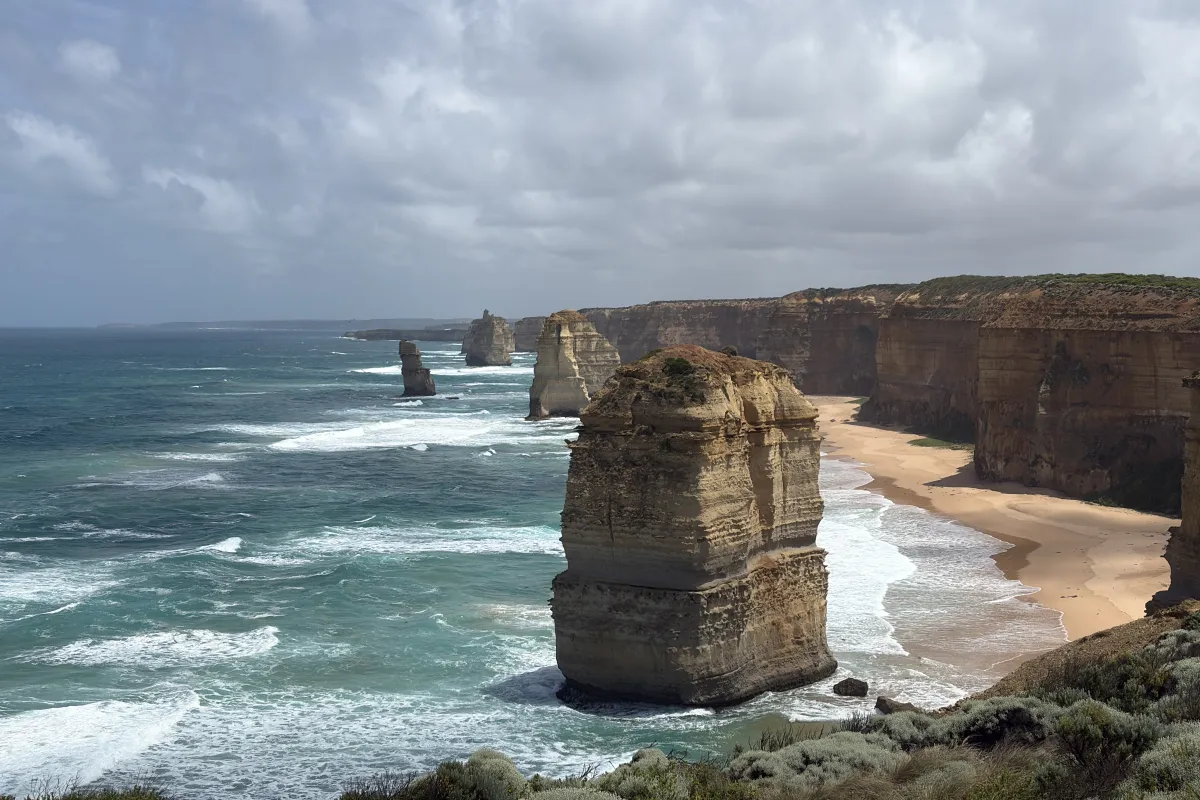 Steile Felsklippen und markante Felsnadeln ragen aus dem Meer entlang einer wilden Küste mit Sandstrand und brechenden Wellen.