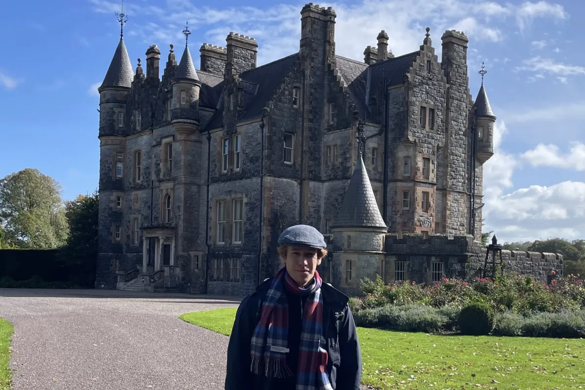Person mit Mütze und Schal steht auf einer Auffahrt vor einem historischen Schloss aus Stein unter blauem Himmel.