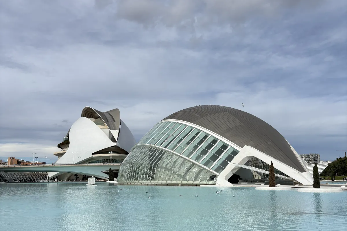 Moderne, futuristische Gebäude mit geschwungenen Glas- und Betonformen an einem großen Wasserbecken unter bewölktem Himmel.