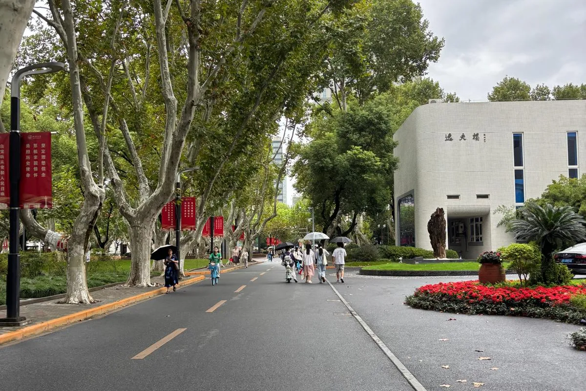 Baumgesäumte Allee auf einem Campus mit Spaziergängern, roten Bannern und einem modernen Gebäude am Straßenrand.