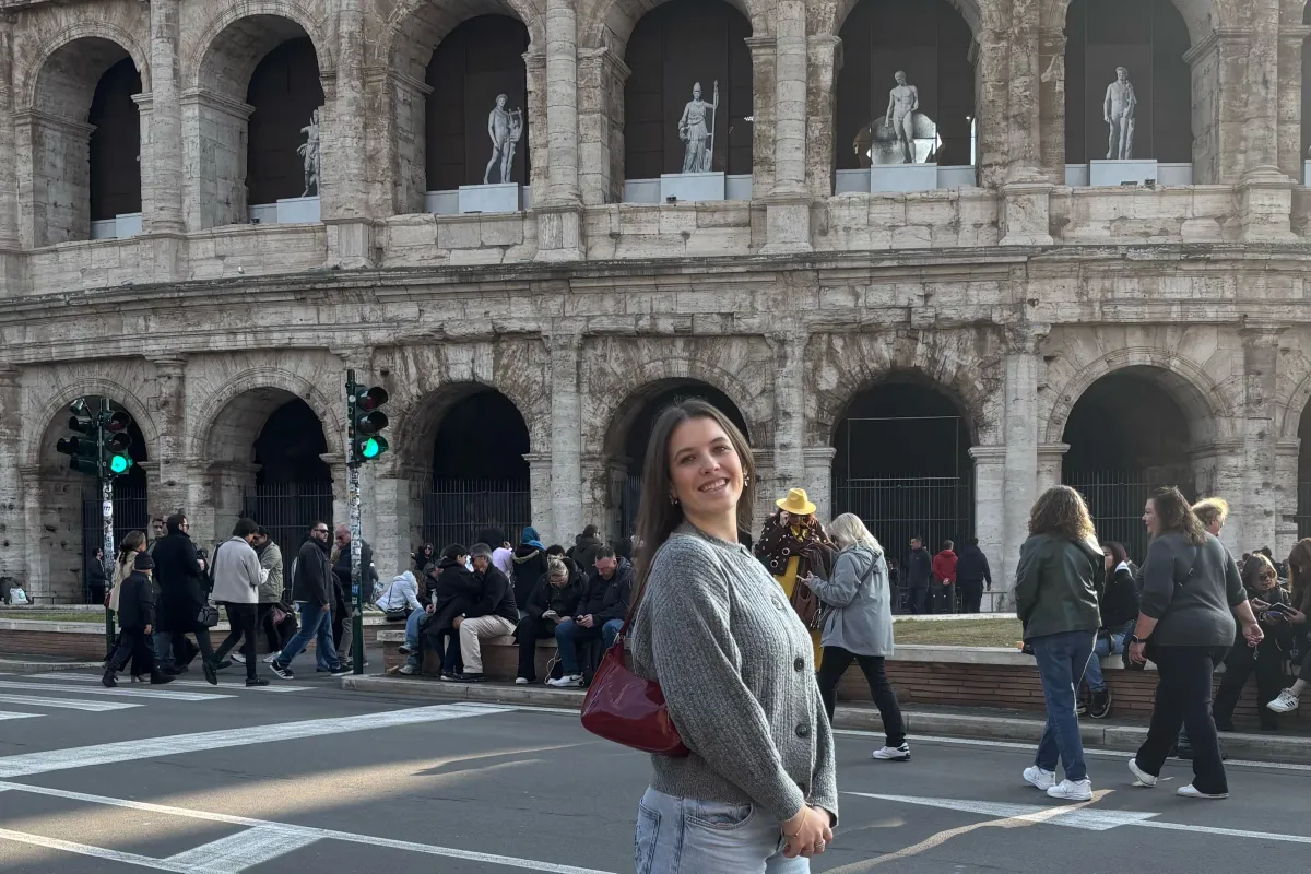 Frau steht lächelnd vor dem Kolosseum, während im Hintergrund Touristen an der Straße entlanggehen.
