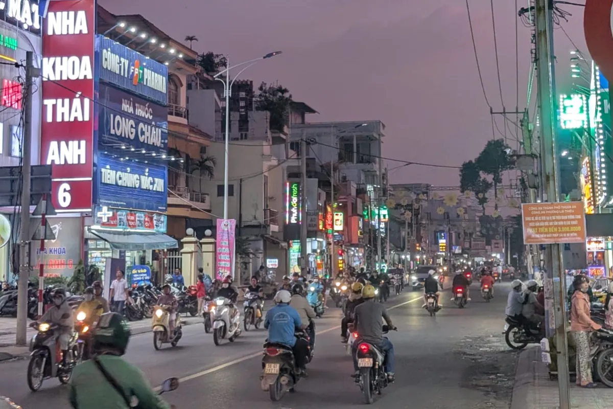 Belebte Straße bei Dämmerung mit vielen Motorrollern, leuchtenden Werbeschildern und dekorativen Lichtern über der Fahrbahn.