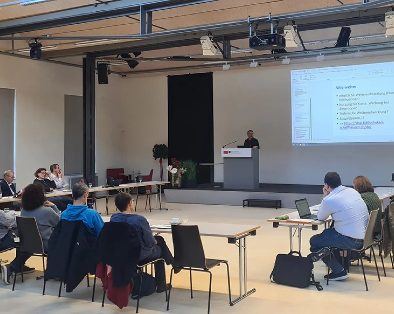 Eine Gruppe von etwa 20 Personen sitzt in einem Auditorium der Universität Liechtenstein, während ein Vortragender an einem Rednerpult spricht. Auf einer großen Leinwand ist eine Präsentation mit dem Titel "Wie weiter" zu sehen.