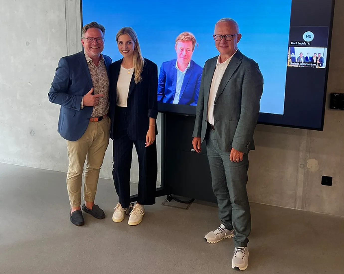 Drei Personen stehen vor einem Bildschirm, der zwei weitere Personen in einem virtuellen Meeting zeigt. Die Szene befindet sich in einem modernen Raum mit Betonwänden.