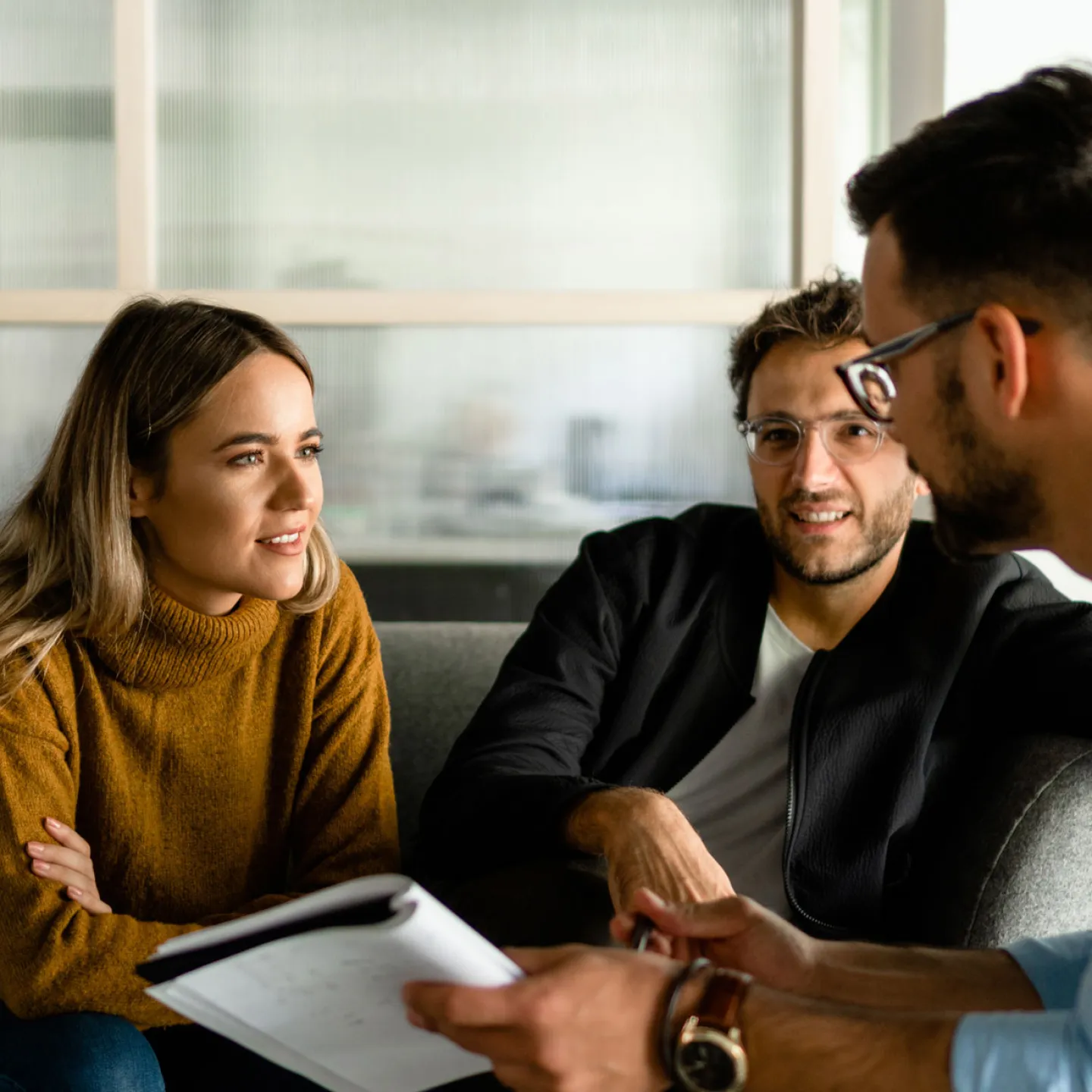 Eine junge Frau und ein junger Mann sitzen nebeneinander auf einem Sofa, während der dritte, der ihnen gegenüber sitzt, sich mit einem aufgeschlagenen Buch in der Hand nach vorne beugt. Der dritte Person führt ein angeregtes Gespräch mit den beiden über das Thema Verantwortung und Gesellschaft. Die konzentrierten Gesichtsausdrücke und die gestikulierende Körpersprache der Gesprächsteilnehmer unterstreichen die Intensität der Diskussion.