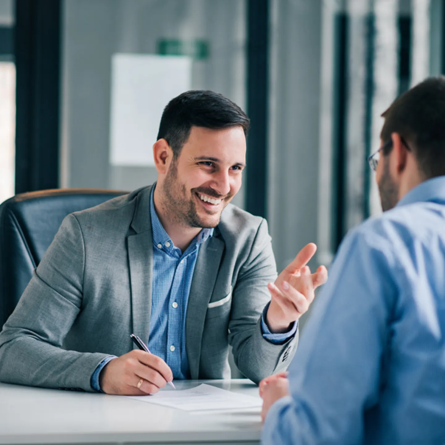 Ein lächelnder Mann in einem grauen Blazer sitzt an einem Schreibtisch und spricht gestikulierend mit einer anderen Person, die ihm gegenüber sitzt. Die Szene vermittelt eine Atmosphäre des professionellen Austauschs und der Beratung.