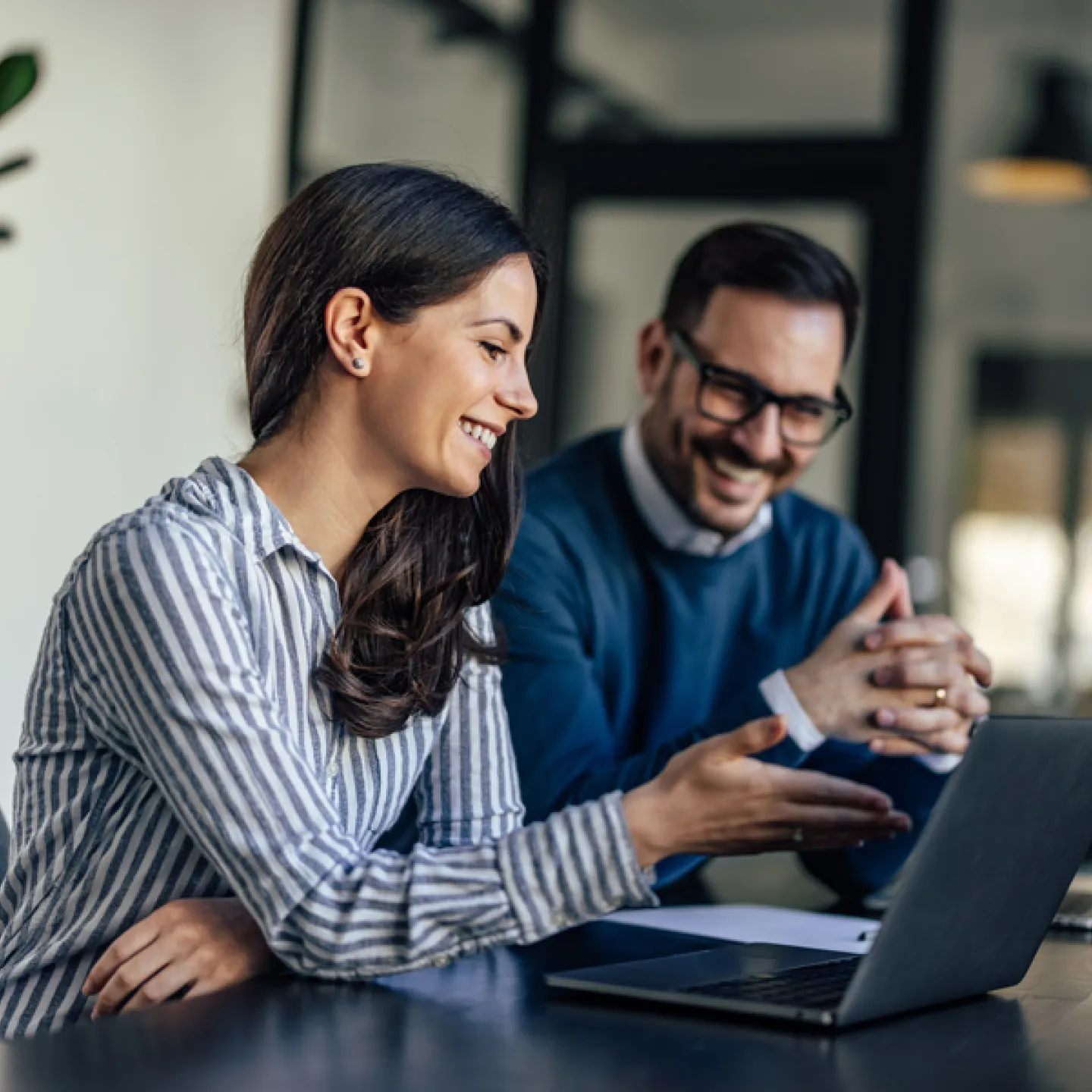 Ein Mann und eine Frau sitzen gemeinsam vor einem Laptop und diskutieren lächelnd über den Bildschirminhalt. Die Atmosphäre wirkt freundlich und kooperativ, während sie sich intensiv mit einer Aufgabe beschäftigen.