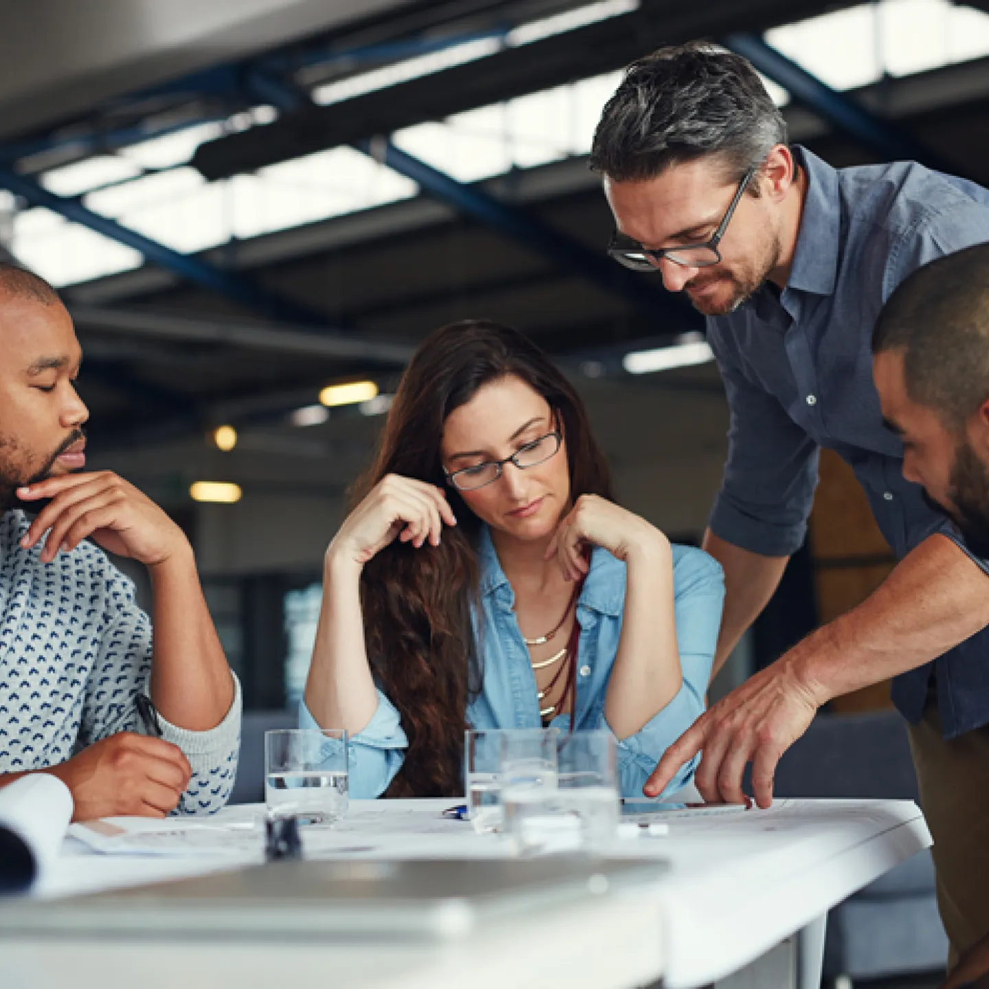 Eine Gruppe von vier Menschen, die um einen Tisch in einem modernen Büro sitzen und gemeinsam an einem Projekt arbeiten. Zwei Männer, eine Frau und ein weiterer Mann, der auf das Dokument zeigt, sind in eine Diskussion vertieft. Vor ihnen liegen Pläne und Dokumente, auf dem Tisch stehen Gläser mit Wasser. Die Atmosphäre wirkt konzentriert und kollaborativ.