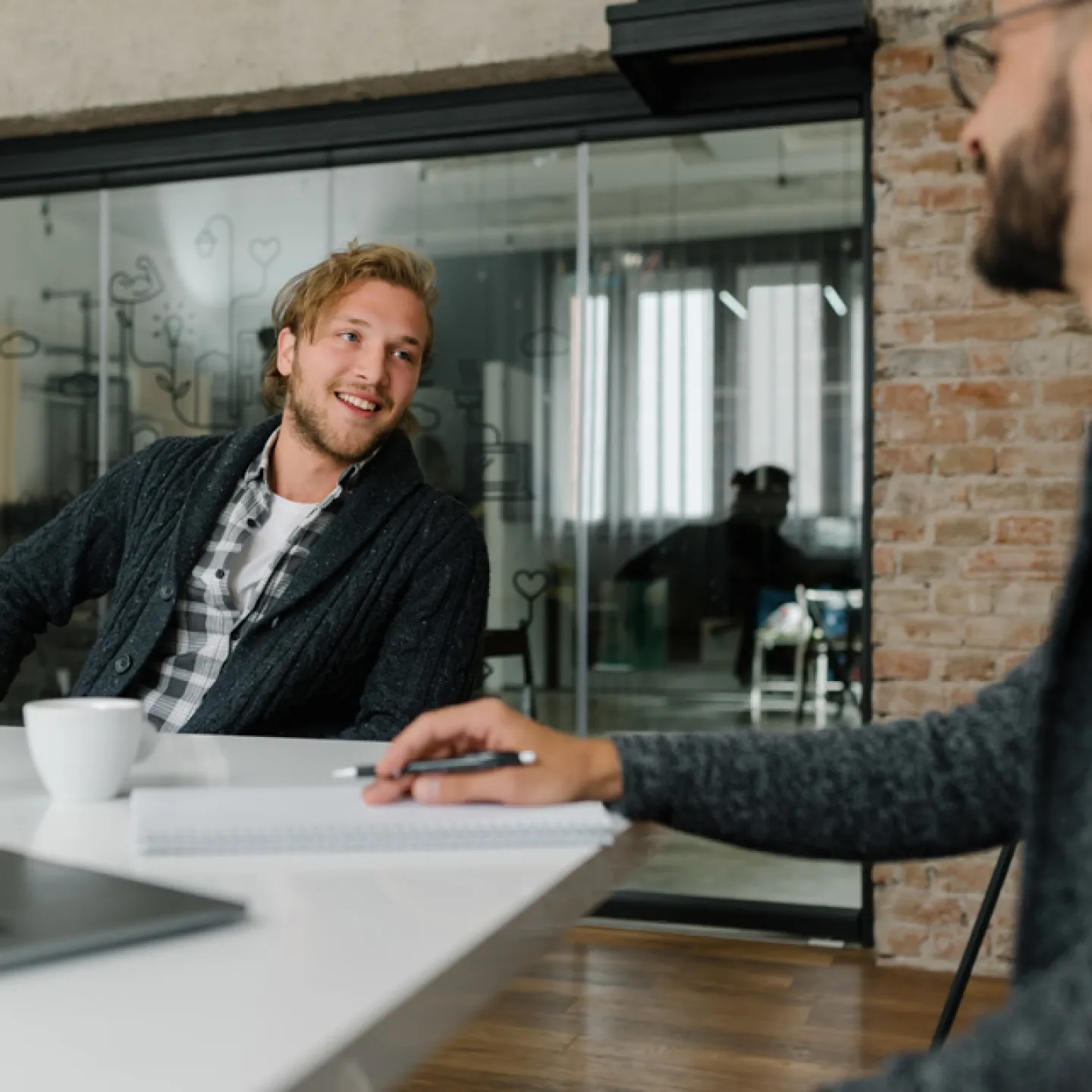 Zwei Männer in einem modernen Büro unterhalten sich an einem Tisch. Der Mann im Vordergrund schreibt in ein Notizbuch, während der andere Mann, der zurückgelehnt und lächelnd ist, zuhört. Auf dem Tisch stehen ein Laptop, ein Glas Wasser und eine Tasse. Im Hintergrund sind Glaswände und Ziegelwände zu sehen, die dem Raum eine offene und zeitgemäße Atmosphäre verleihen.