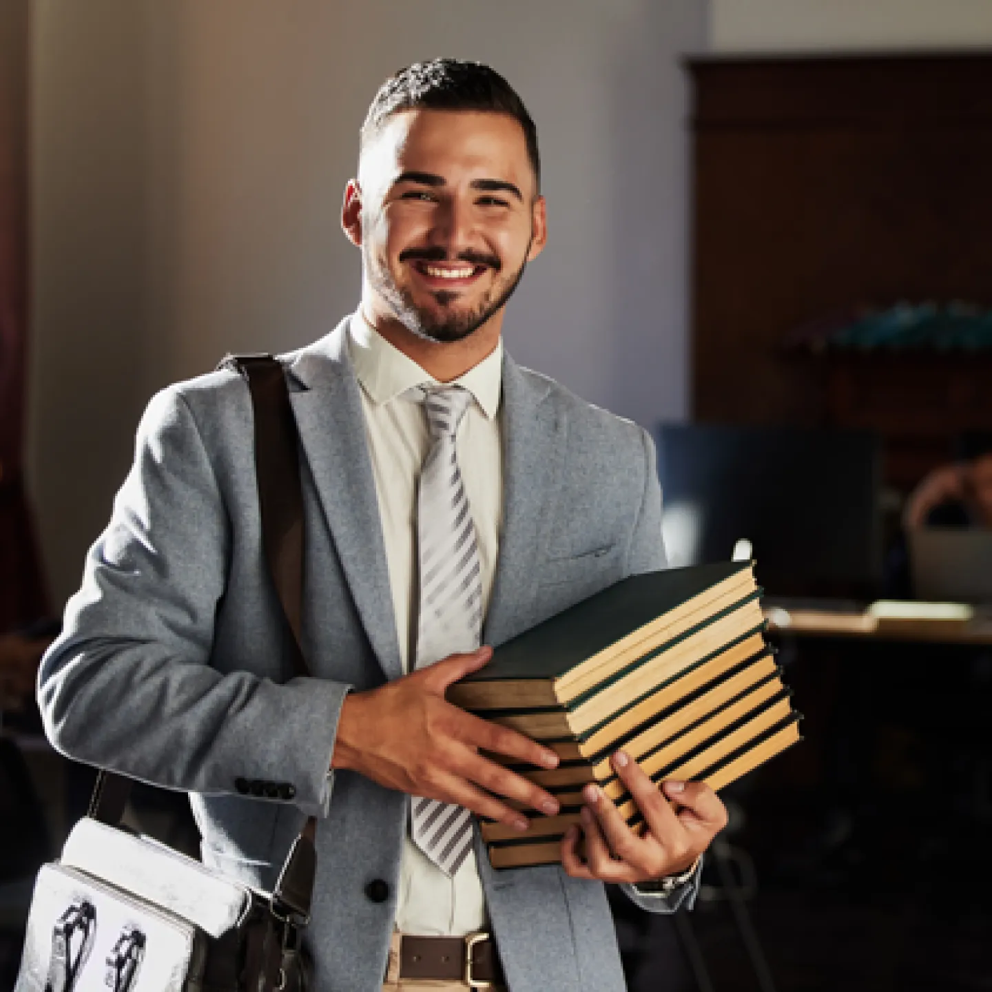 Ein junger, selbstbewusster Mann in einem Business-Anzug, der mehrere Bücher trägt, lächelt freundlich in die Kamera. Der Hintergrund zeigt eine ruhige Büroumgebung, die eine produktive und professionelle Atmosphäre vermittelt.