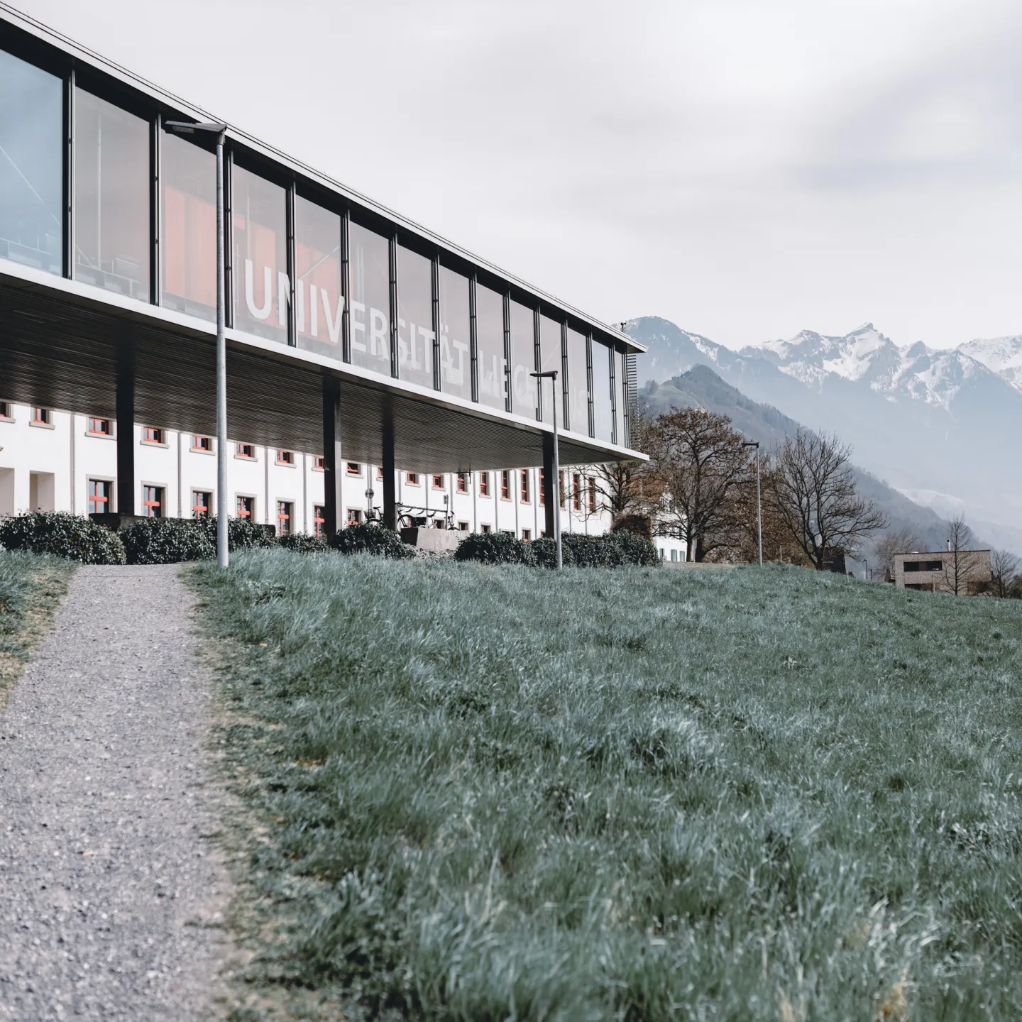 Campus Liechtenstein mit Glasfassade, Wiesenweg und Panoramablick auf die umliegenden Berge und das Dorf Vaduz.