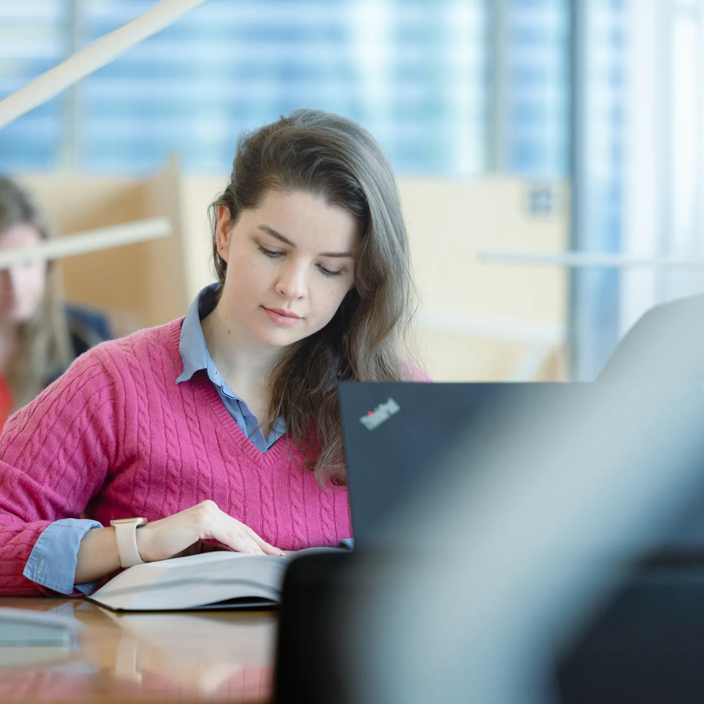 Studentin der Universität Liechtenstein arbeitet konzentriert mit Buch und Laptop in der lichtdurchfluteten Bibliothek.